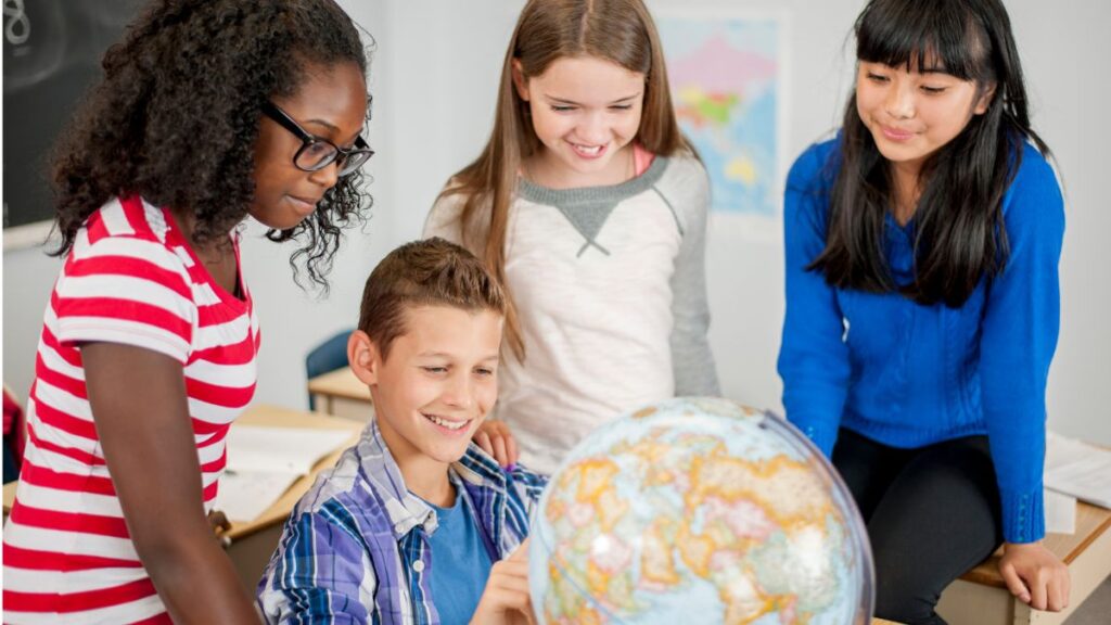 Students aged around 10 to 14 pointing at a world map in a bright classroom and learning what is social studies.